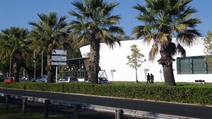 Un bâtiment moderne, avec des palmiers devant, une enseigne indiquant « Couchant Petite Motte Les Plages », et une route où circulent des voitures et des piétons.