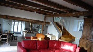 A spacious living area with a red leather sofa, wooden beams on the ceiling, a staircase, and a kitchen in the background. - House, charming cottage in the Cantal (Bassignac)