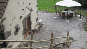 A rustic stone building with a gravel patio, a wooden fence, and a white umbrella table with chairs. - House, charming cottage in the Cantal (Bassignac)