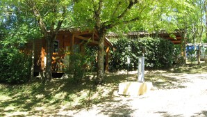 A rustic wooden cabin surrounded by lush greenery, with a covered porch and a stone pathway leading to it. - Camping Le Faillal - Cabanes et chalets (Montpezat-de-Quercy)