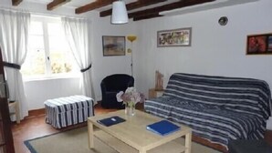 Una habitació acollidora amb un llit cobert per un llençol a ratlles, una taula petita amb un ram i llibres, i una finestra amb cortines.