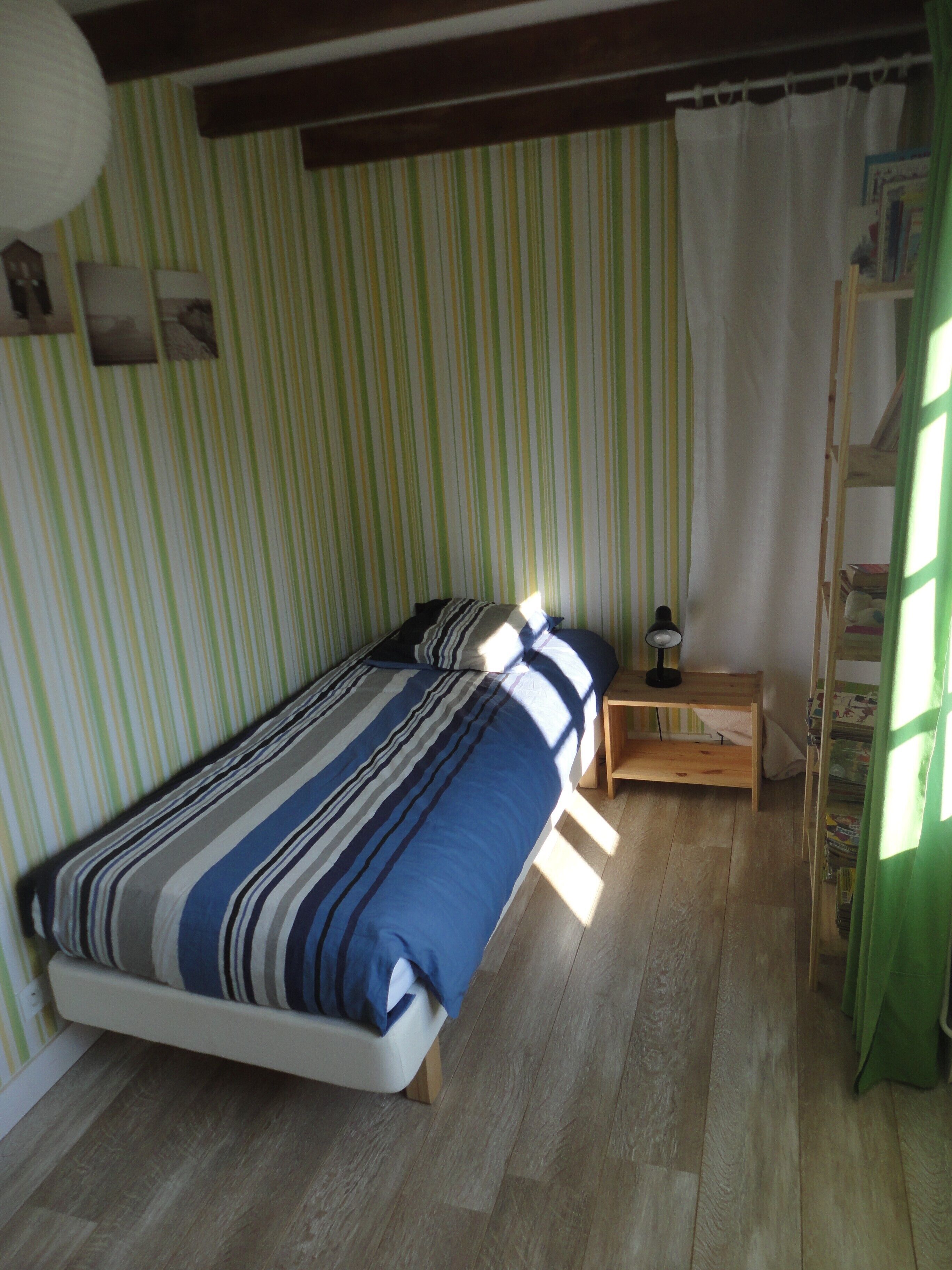 Uma cama de solteiro com colcha listrada, uma mesa de cabeceira com luminária e uma escada de madeira encostada na parede.