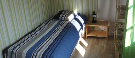 Uma cama de solteiro com colcha listrada, uma mesa de cabeceira com luminária e uma escada de madeira encostada na parede.