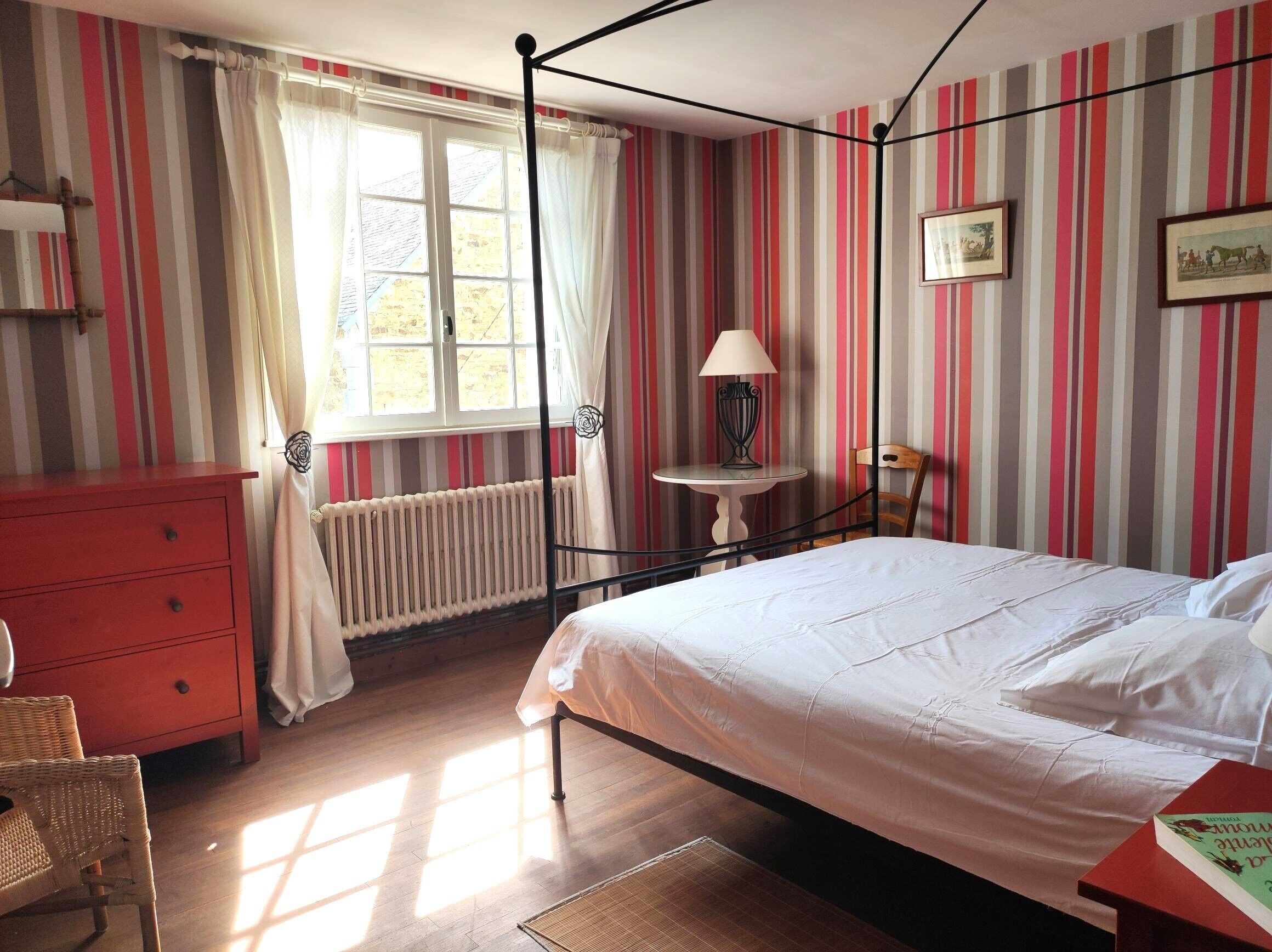 Una habitación con una cama con dosel, un aparador rojo, una mesita con una lámpara y una silla.
