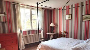 Habitació amb un llit amb cortines, un aparador vermell, una taula petita amb una làmpada i una cadira.
