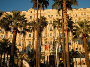 A large, historic building with multiple balconies and flags, surrounded by palm trees. - Air-conditioned 3-room apartment, historic town center, 200m from the beach (Cannes)