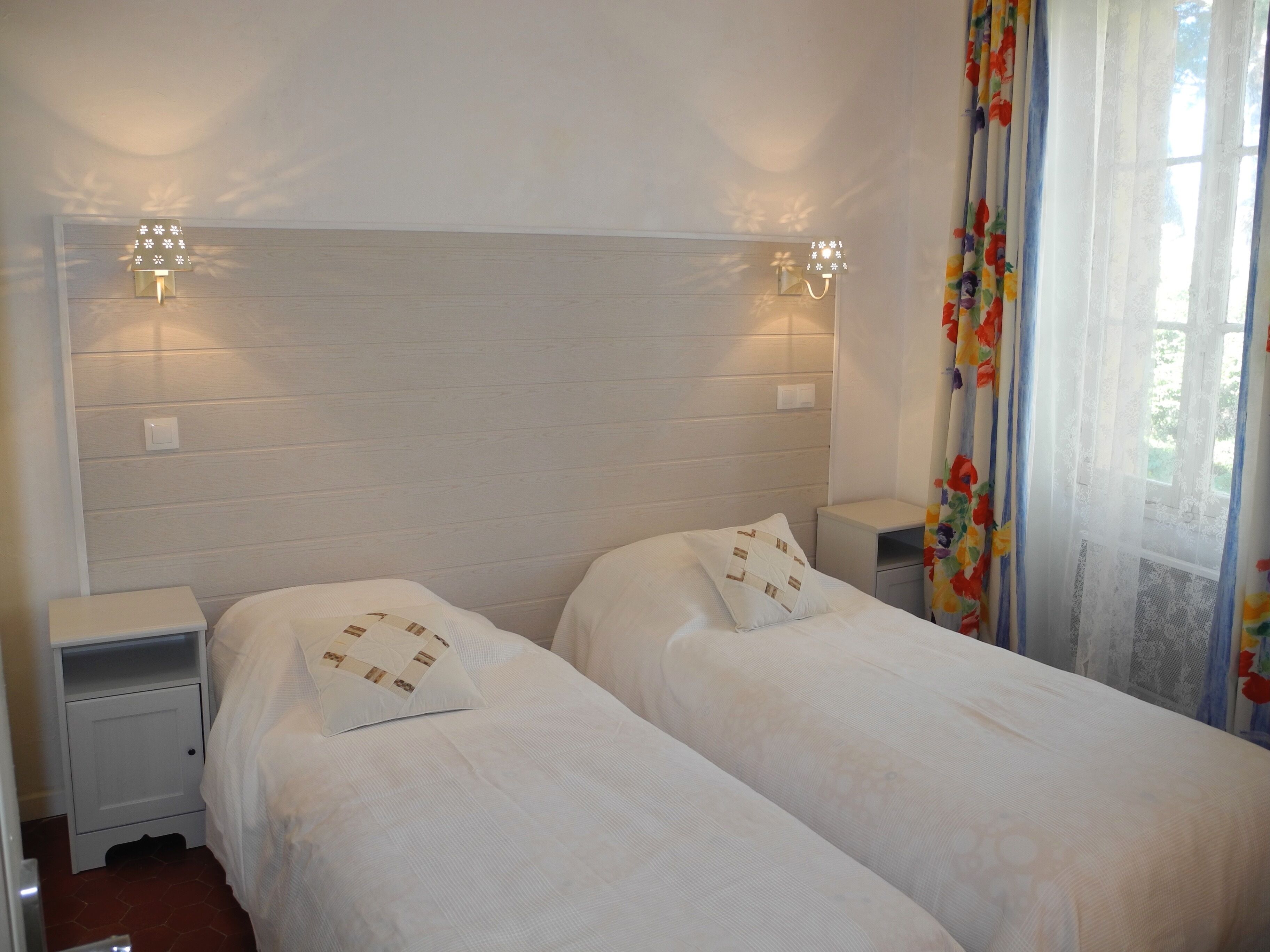 Ein Doppelbett mit gestepptem Kopfkissen, ein Nachttisch, eine Wandleuchte und ein Fenster mit floralem Vorhang.