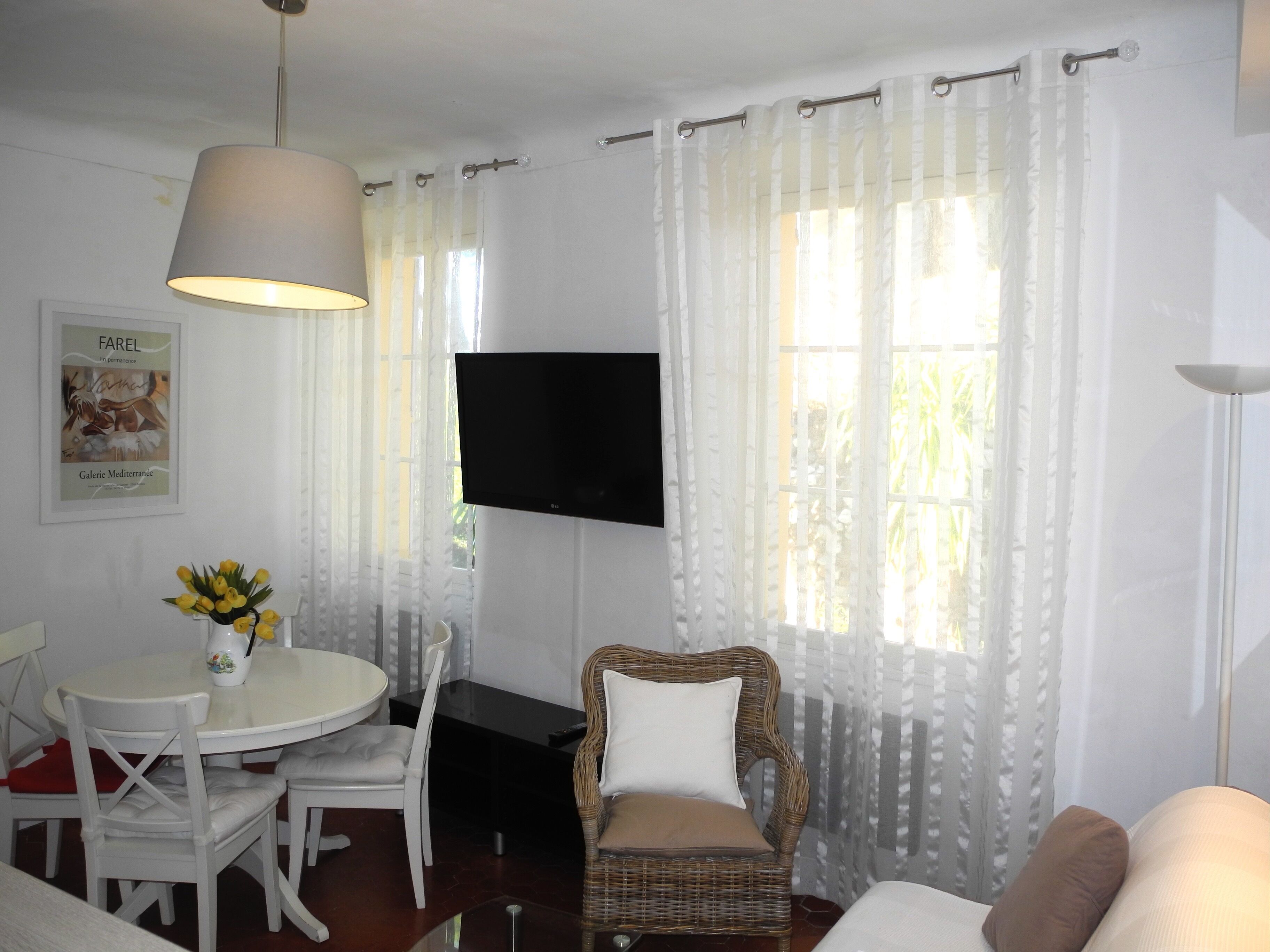 A living room with a dining area, a flat-screen TV mounted on the wall, and a wicker chair.