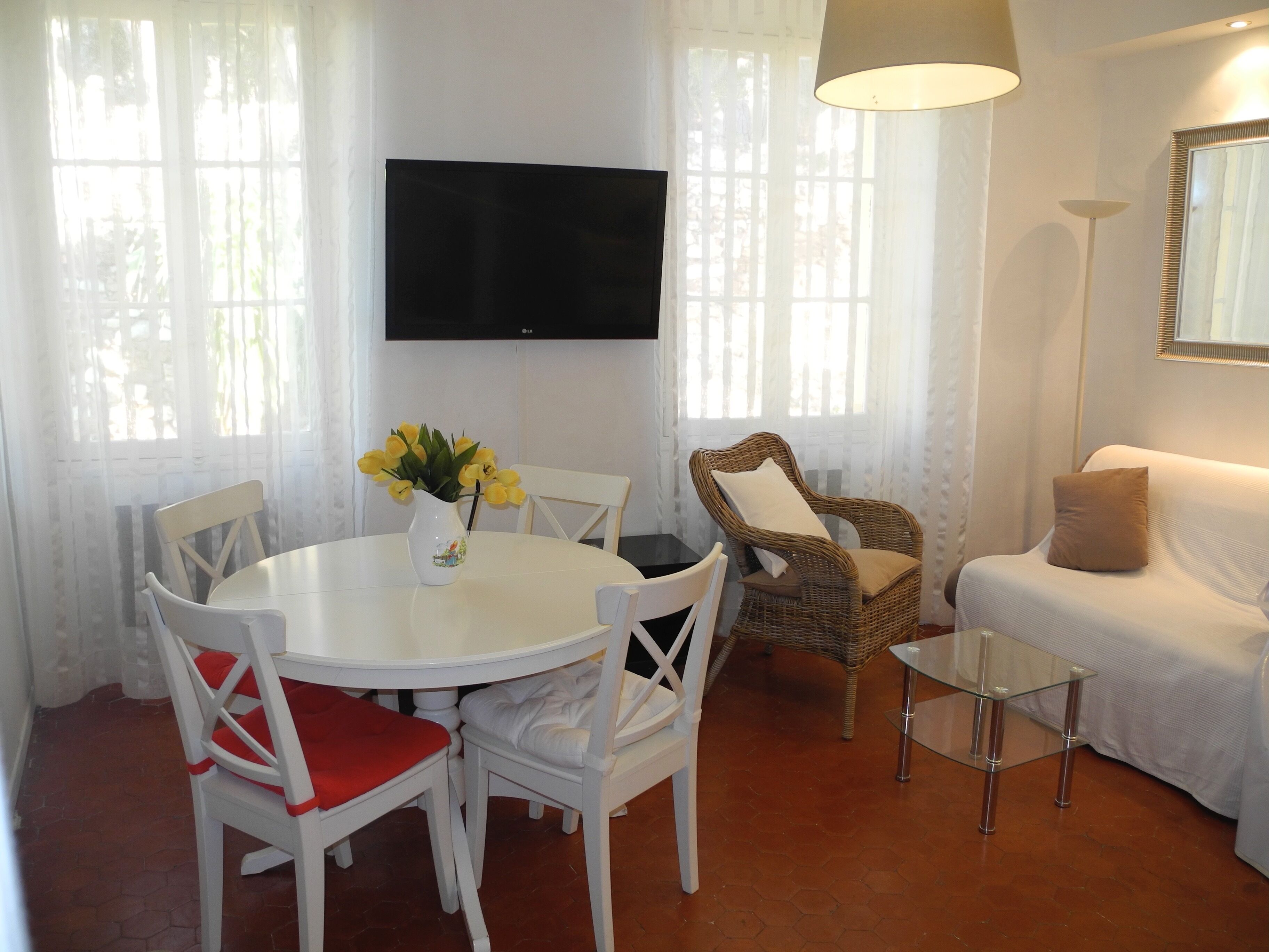 A living room with a round dining table, white chairs, a wicker chair, a sofa, a glass coffee table, and a TV mounted on the wall.