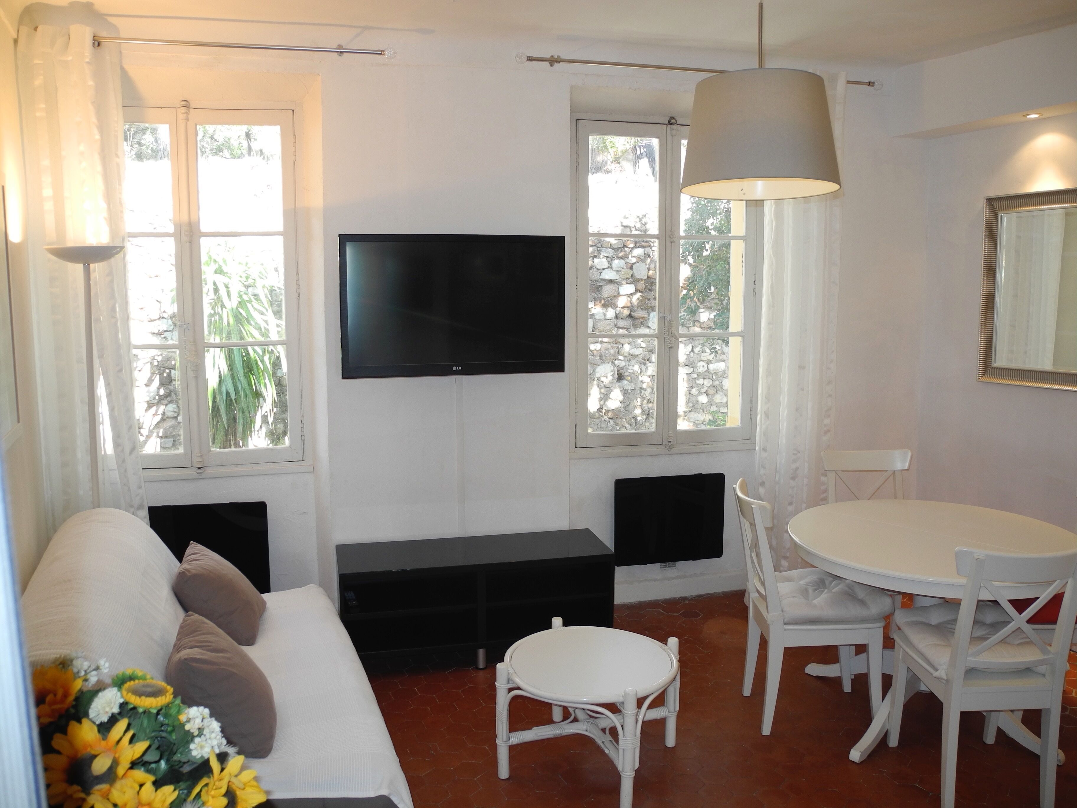 A room with a white sofa, a television mounted on the wall, a dining table with chairs, and a window with a view of greenery.