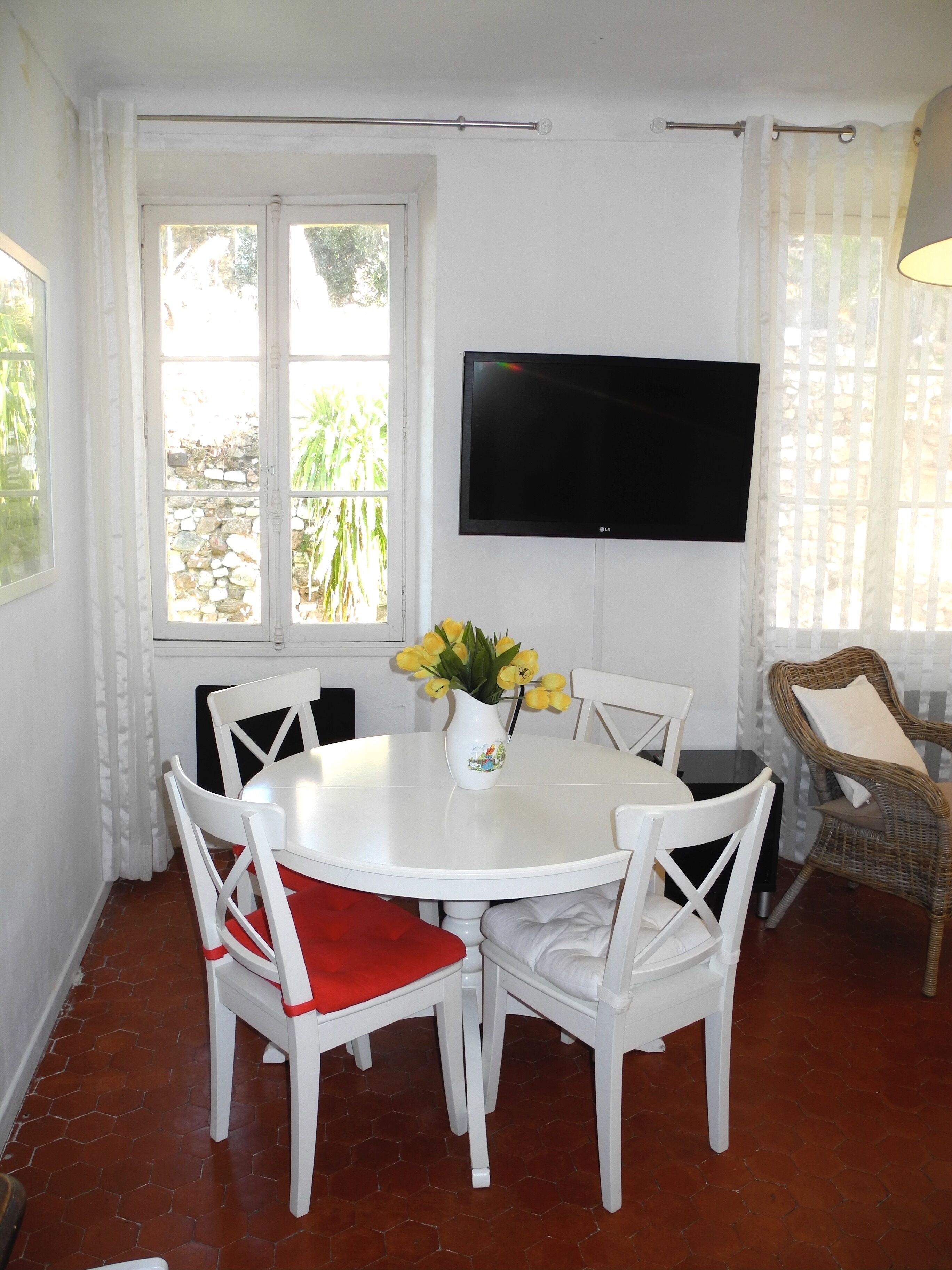 A dining area with a white table and chairs, a vase with yellow flowers, a flat-screen TV on the wall, and a wicker chair with a cushion.