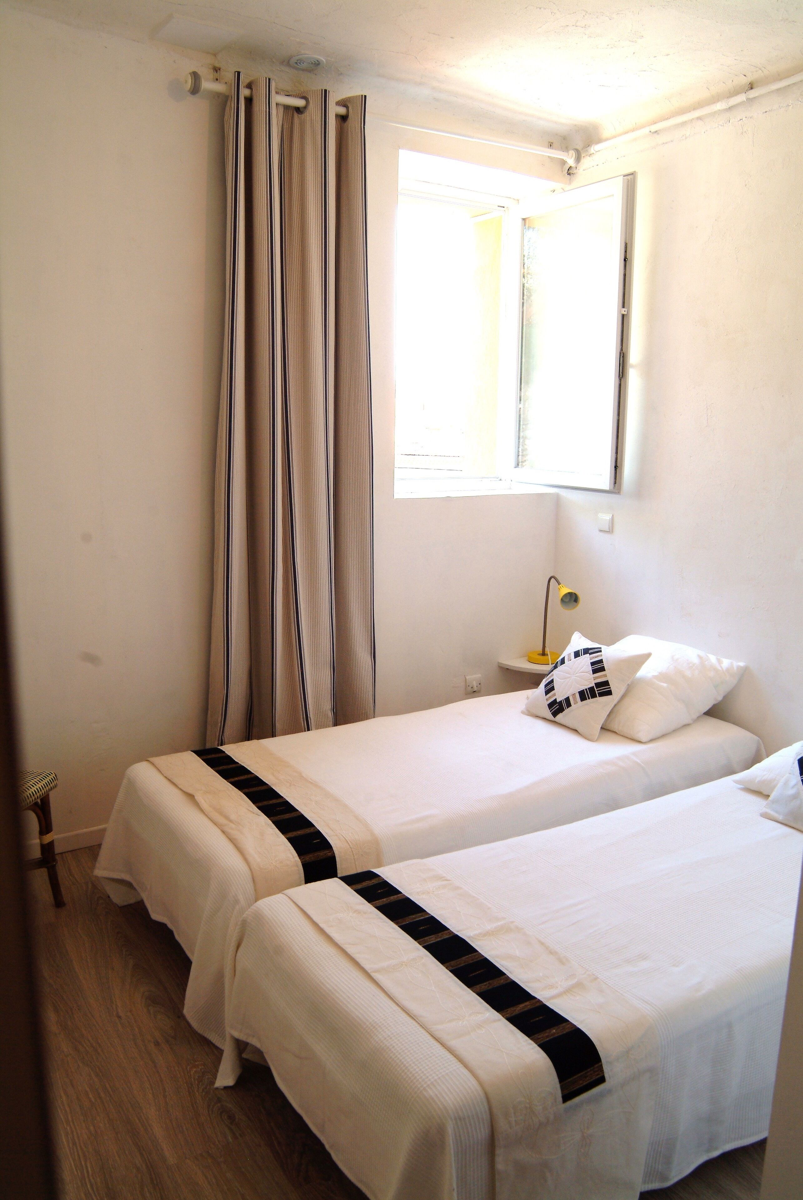 Una habitación con dos camas, una ventana con cortinas, una lámpara de noche y una mesita.