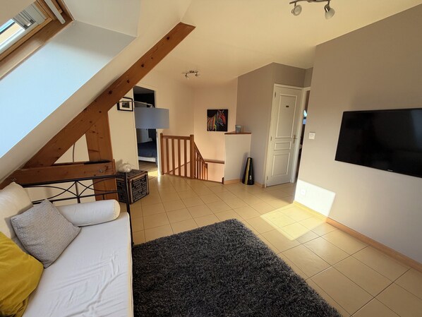 A loft-style room with a sloped ceiling, a sofa, a TV, and a staircase leading to another room. - Luxury contemporary villa 300 m from the sea – 10 people (Saint-Germain-Sur-Ay)