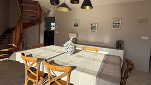 A dining area with a large table, wooden chairs, pendant lights, a patterned tablecloth, and a staircase.