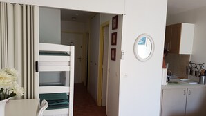 A room with bunk beds, a dining table with flowers, a kitchen with cabinets, and a hallway leading to another room. - Le Lavandou, La Fossette appt 2/4 pers. beach at 150m. (Le Lavandou)