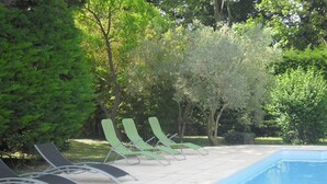 En swimmingpool med ett kaklat poolområde, omgiven av gröna träd och två solstolar.