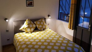Quarto com cama, duas luminárias de cabeceira, uma janela com cortinas e uma pequena imagem na parede.