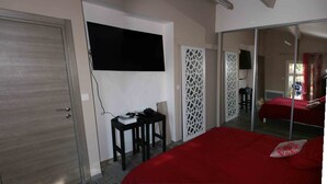 Un dormitorio con una cama grande, un escritorio con silla, un televisor montado en la pared y una puerta corredera que da a otra habitación.