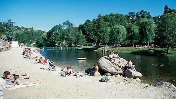 A riverside beach with people relaxing on the sand and rocks, a boat on the water, and trees in the background.