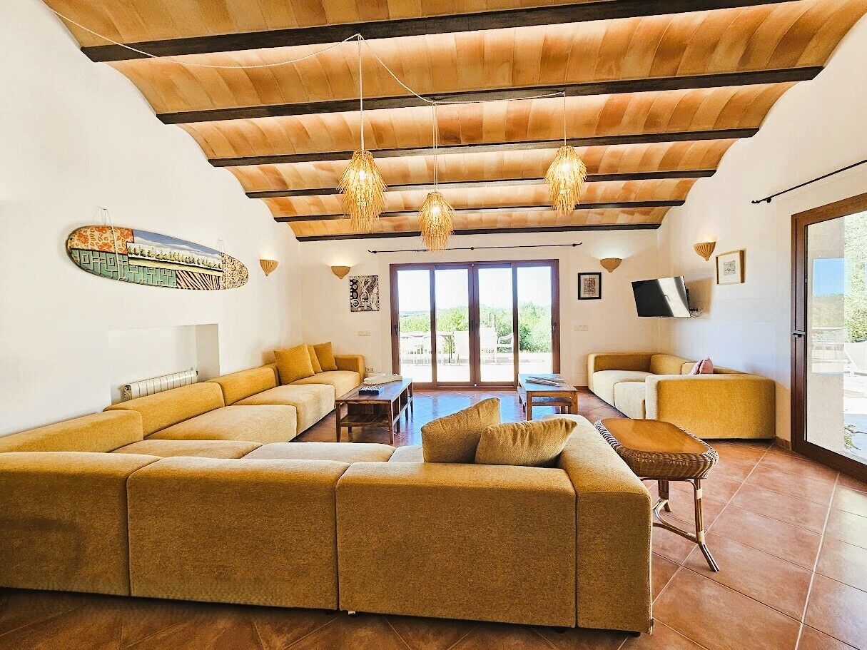 Un salon spacieux avec un grand canapé d’angle, une table basse et un coin repas. Des luminaires suspendus et une grande fenêtre laissent entrer la lumière naturelle.