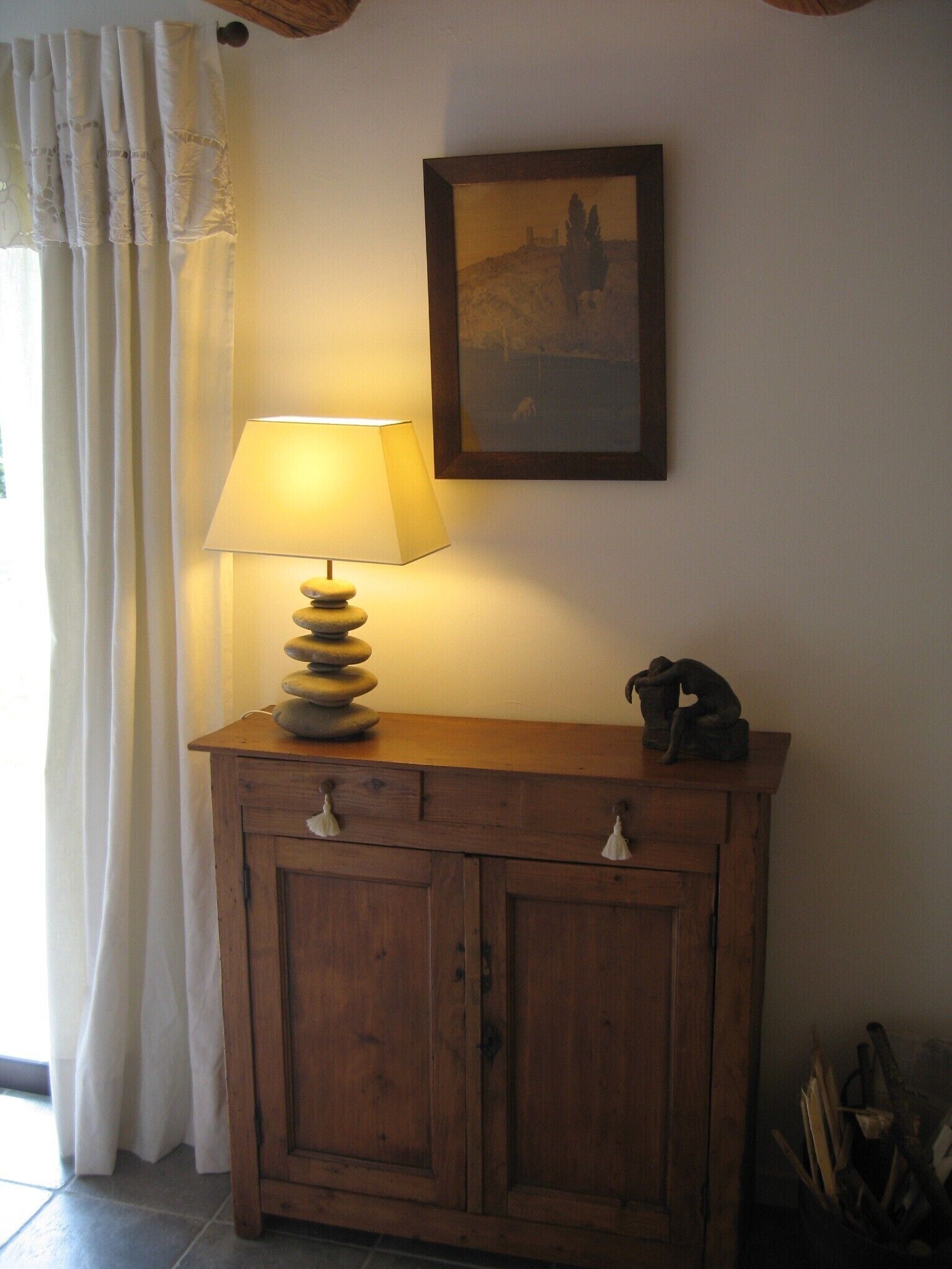 Un meuble en bois surmonté d’une lampe, un tableau encadré accroché au mur et un rideau tiré partiellement sur le côté.