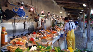 Un mercado de alimentos con diversos productos expuestos en cestas y recipientes, una caja registradora y una persona con delantal.