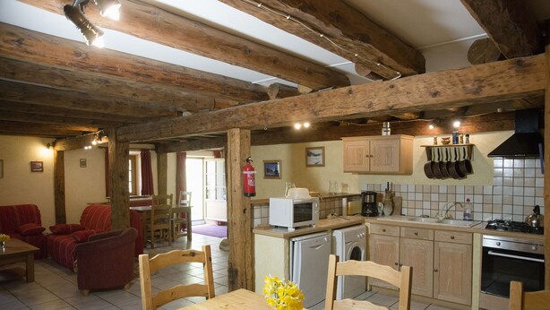 A rustic kitchen with wooden beams, a dining area with a wooden table and chairs, and a living room with red sofas. - Renovated 120 square metre gîte in old farmhouse (La Pesse)