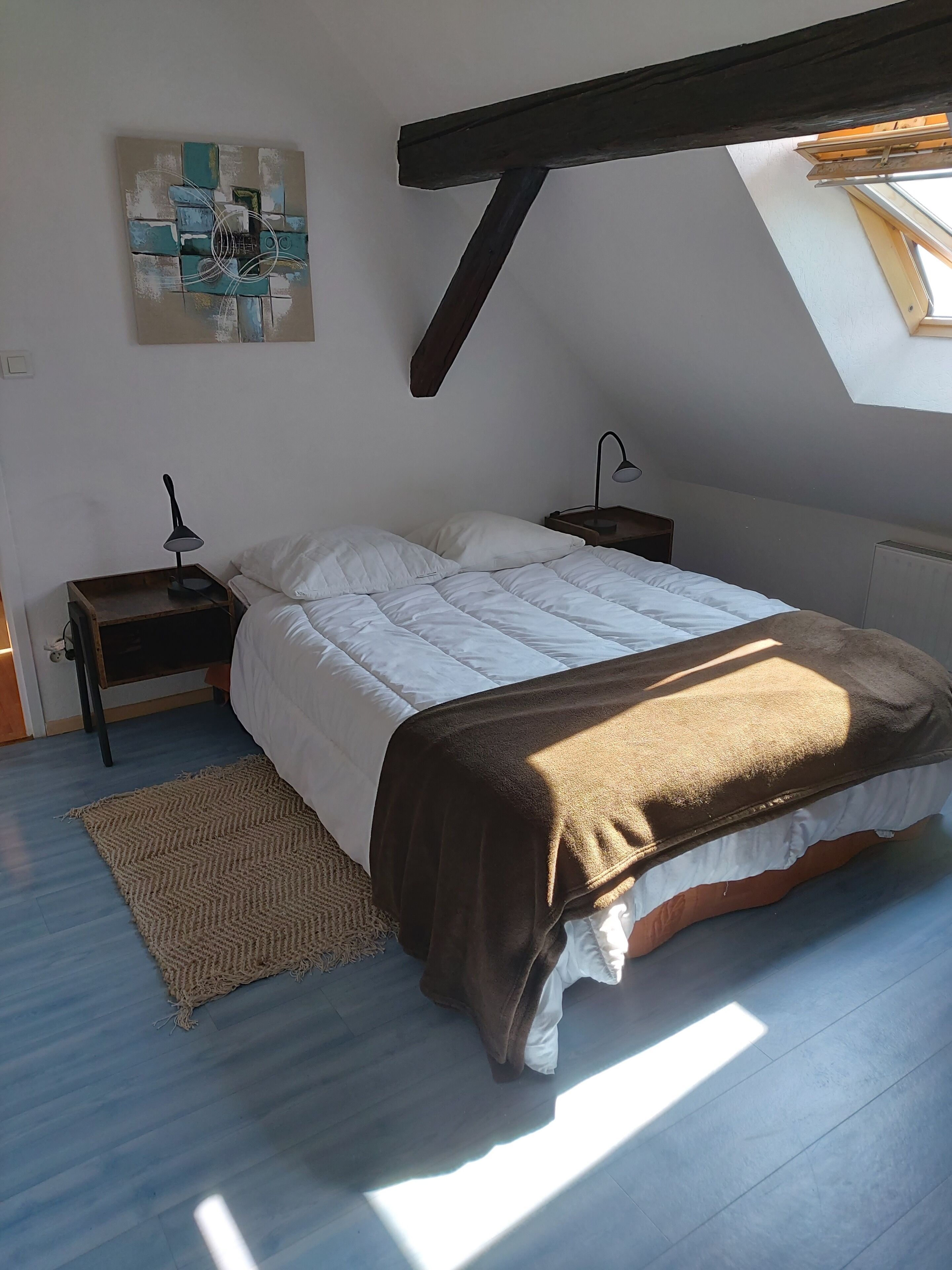 A bedroom with a skylight, a bed with a brown blanket, a nightstand with a lamp, and a painting on the wall.