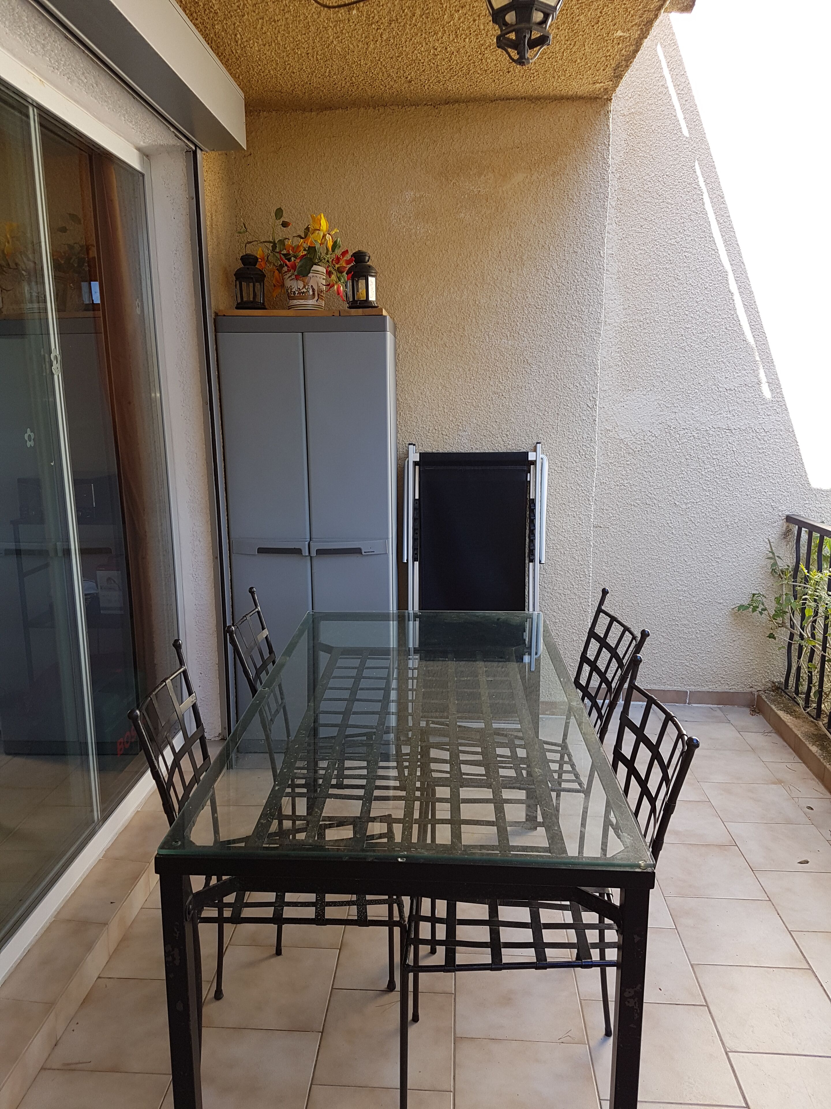 Uma mesa com tampo de vidro, quatro cadeiras, um armário cinzento e uma planta pequena numa varanda com soalho em azulejos.