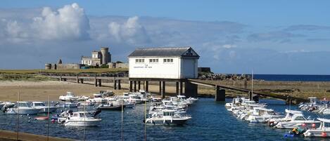 A coastal marina with multiple boats docked, a bridge leading to a white building with signage, and a historic stone structure in the background.