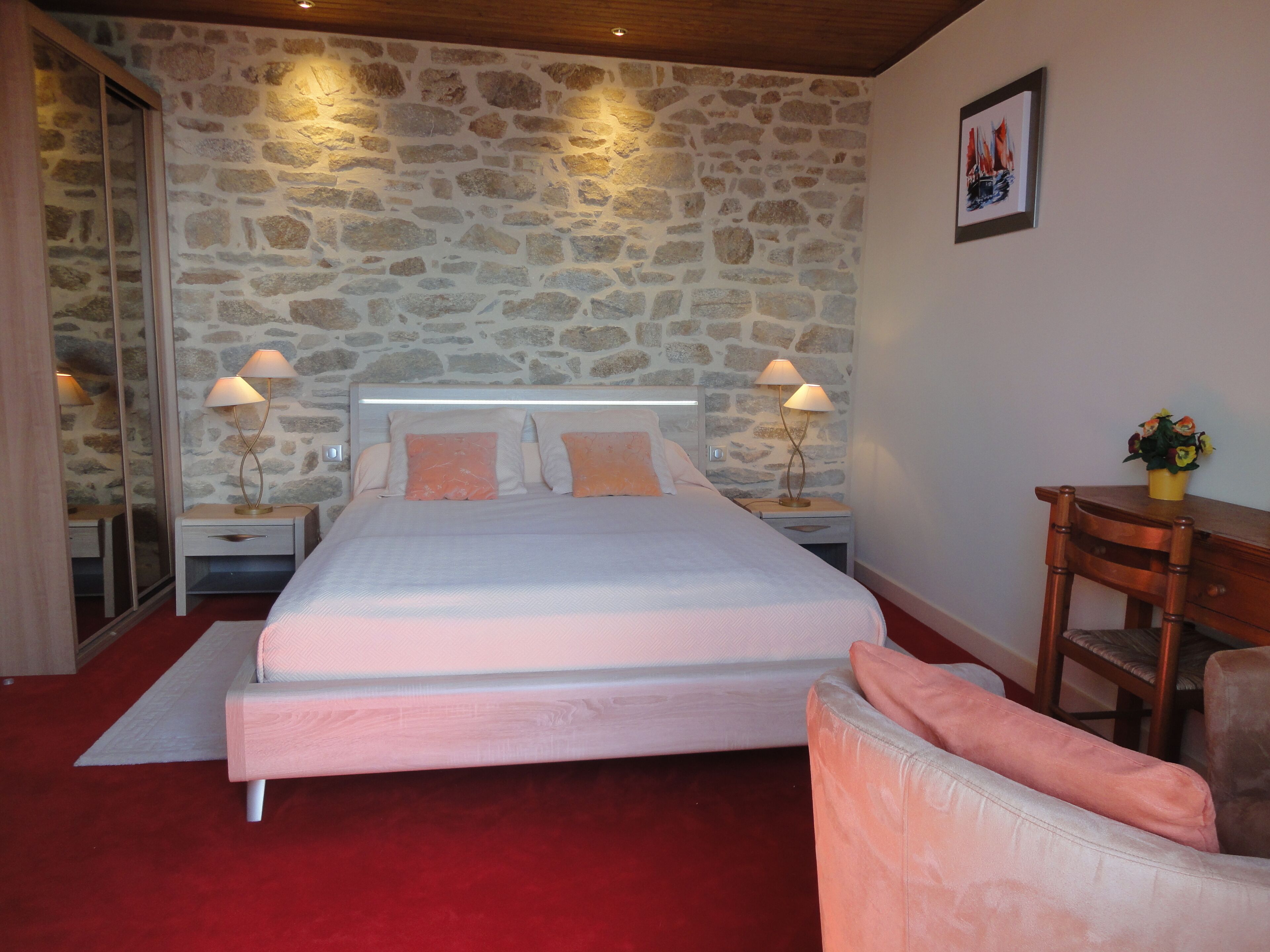 A bedroom with a stone wall, a bed with two pillows, two bedside tables with lamps, a wooden chair, and a small table with a vase.