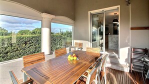 A spacious balcony with a wooden table and chairs, a glass door leading to a kitchen, and a view of a hedge and sky. - Beautiful Villa with Large Heated Pool and Sea Water, 5 km from the Beach (Portiragnes)