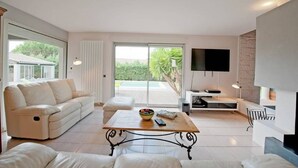 A modern living room with a sofa, a coffee table, a TV, and a view of a pool and greenery outside. - Beautiful Villa with Large Heated Pool and Sea Water, 5 km from the Beach (Portiragnes)