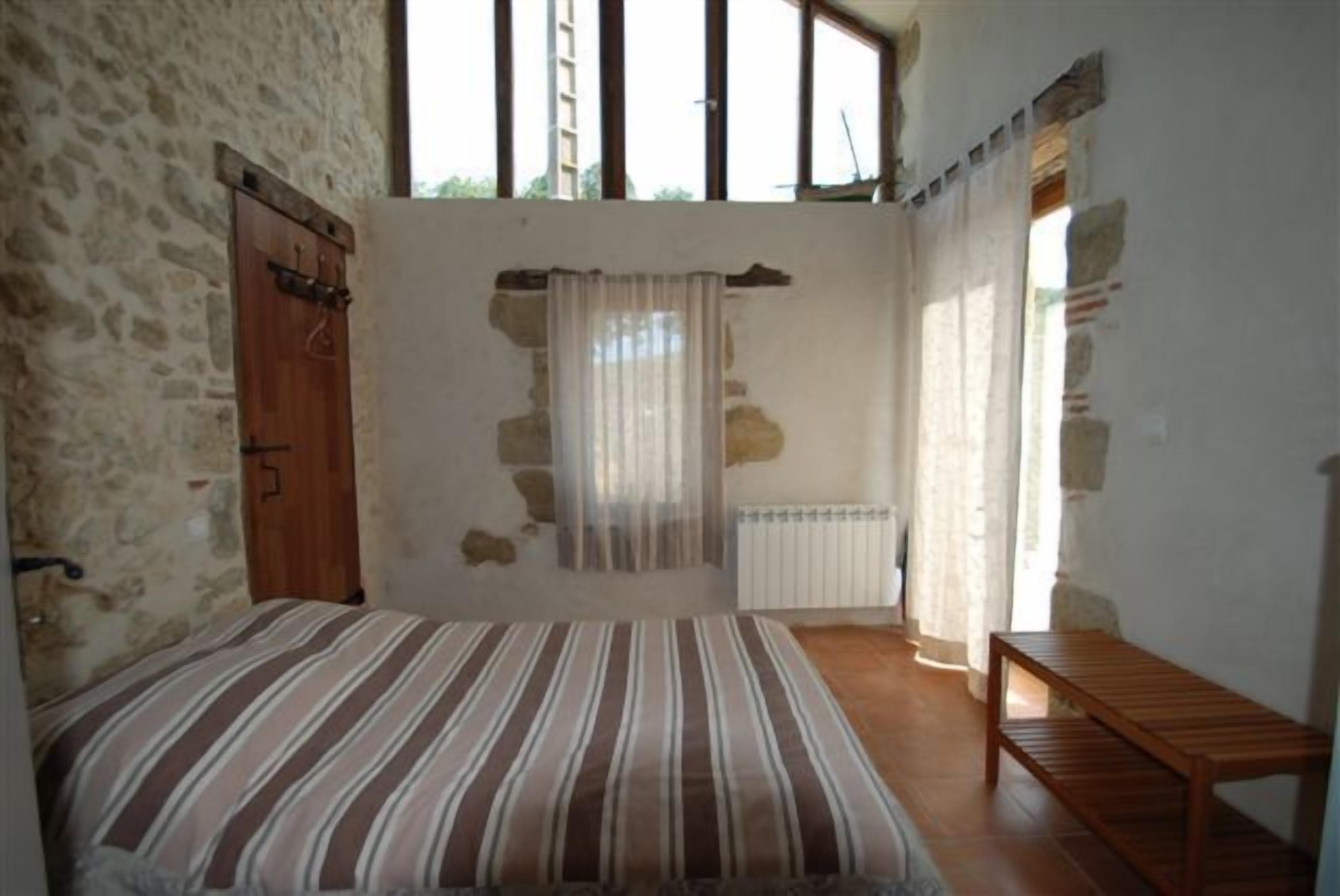 A bedroom with a bed, wooden bench, stone walls, and large windows.