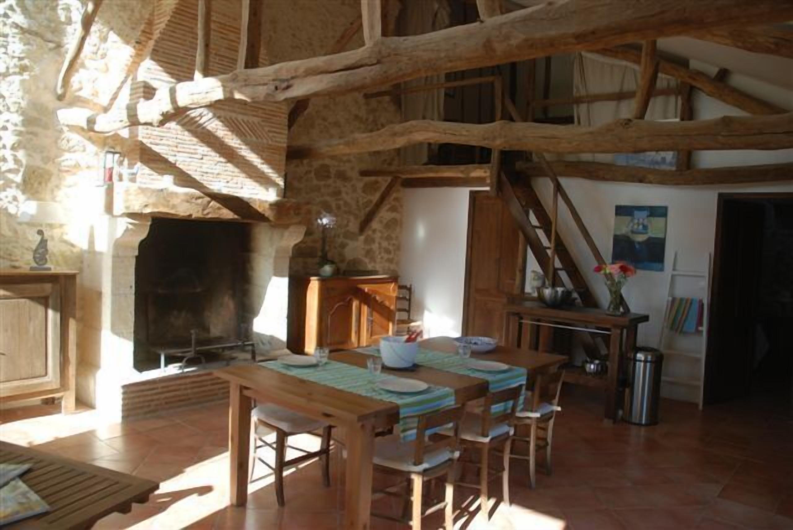Un coin repas rustique avec une table en bois dressée pour quatre personnes, une cheminée et un escalier menant à un niveau supérieur.