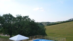 A wooden deck with a pool, a covered seating area, and a view of a green landscape.