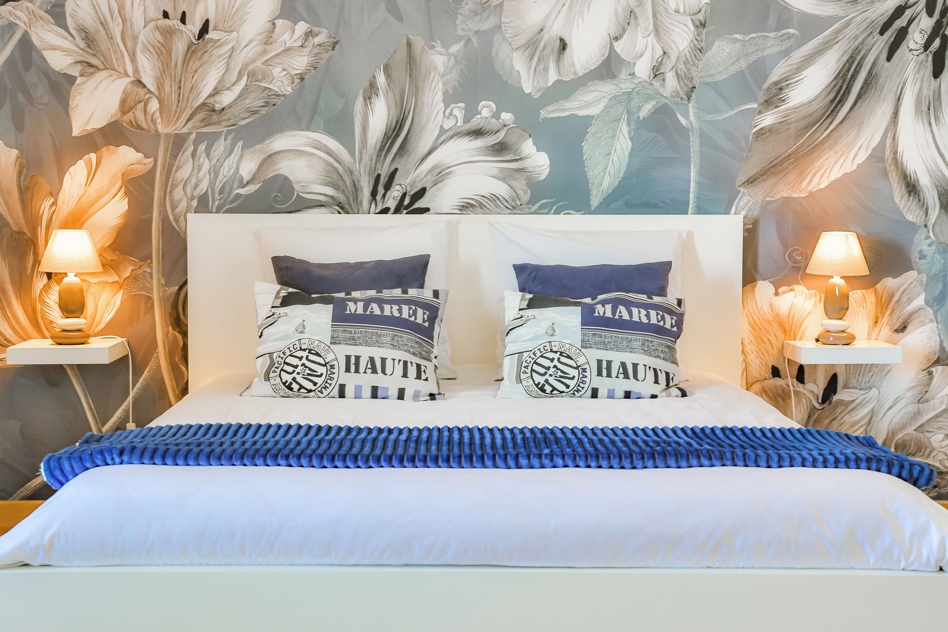 A neatly made bed with a blue accent, two matching pillows, and a floral wallpaper backdrop.