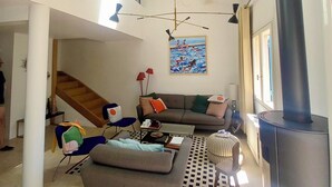 A spacious living area with a high ceiling, a large black stove, a sofa set, a dining table, and a staircase leading to an upper level.
