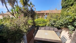 A patio with a wooden table and chairs, surrounded by lush greenery and a brick wall.