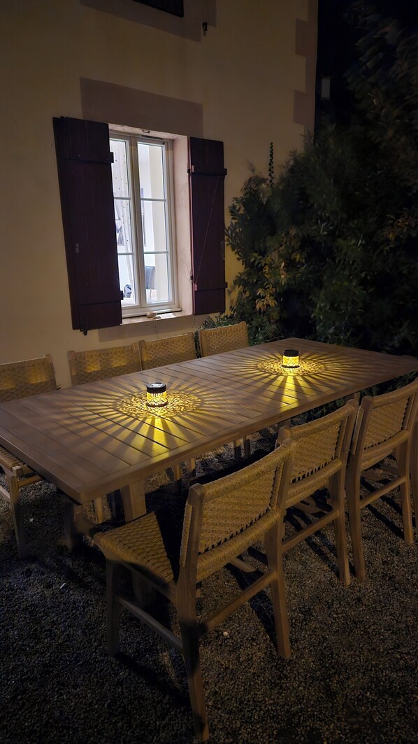 An outdoor dining area with a table and chairs, a window with open shutters, and a view of greenery.