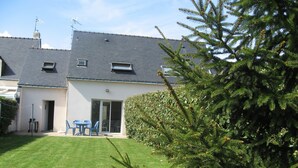 A two-story house with a sloped roof, a small garden, and a blue table and chairs outside.