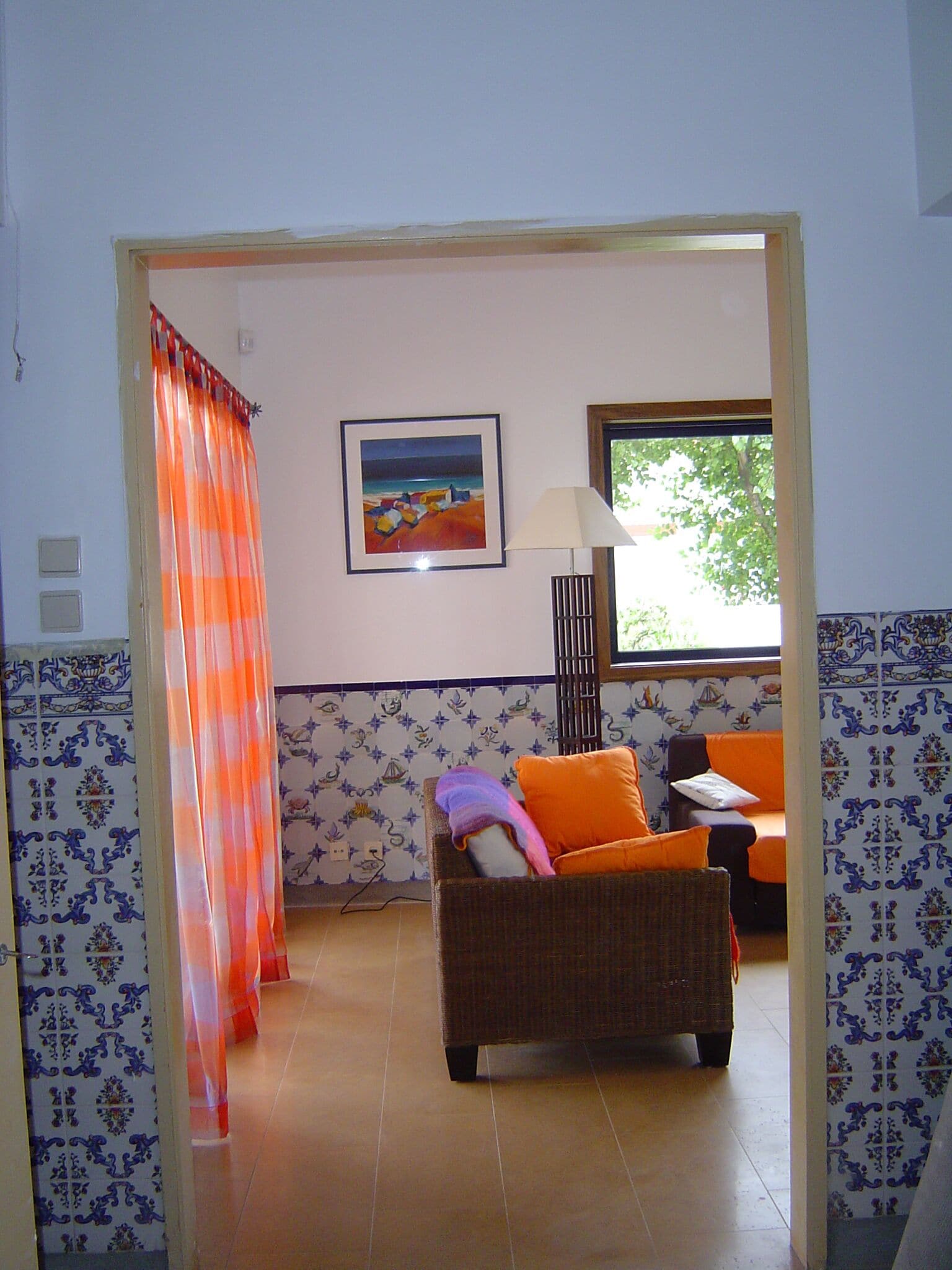 Une pièce avec un canapé en osier, un coussin orange, un tableau encadré et une fenêtre donnant sur un paysage verdoyant.