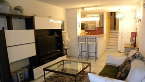 Sala de estar com televisão, sofá e mesa de centro. Cozinha ao fundo, com pia e armários.