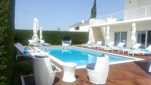 Una piscina con sillas de descanso y sombrillas, rodeada por una terraza de madera y un seto.