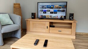 Un salón con una televisión de pantalla plana sobre un soporte de madera, dos controles remotos en una mesa de centro y un sofá gris.
