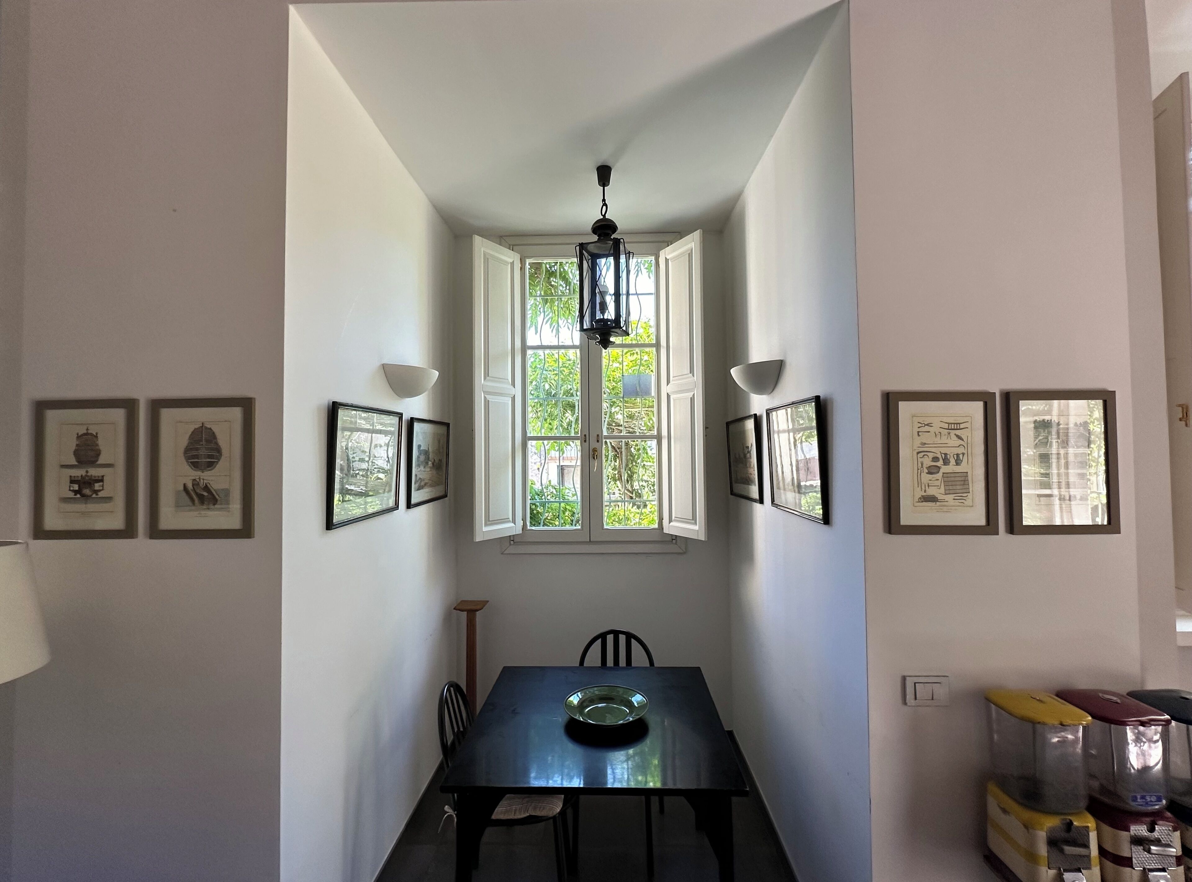 Um corredor estreito com uma mesa de jantar no final, quadros nas paredes e uma janela com vista para o verde.