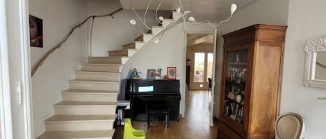 A spacious living room with a wooden staircase, a chandelier, a piano, and a large wooden cabinet.