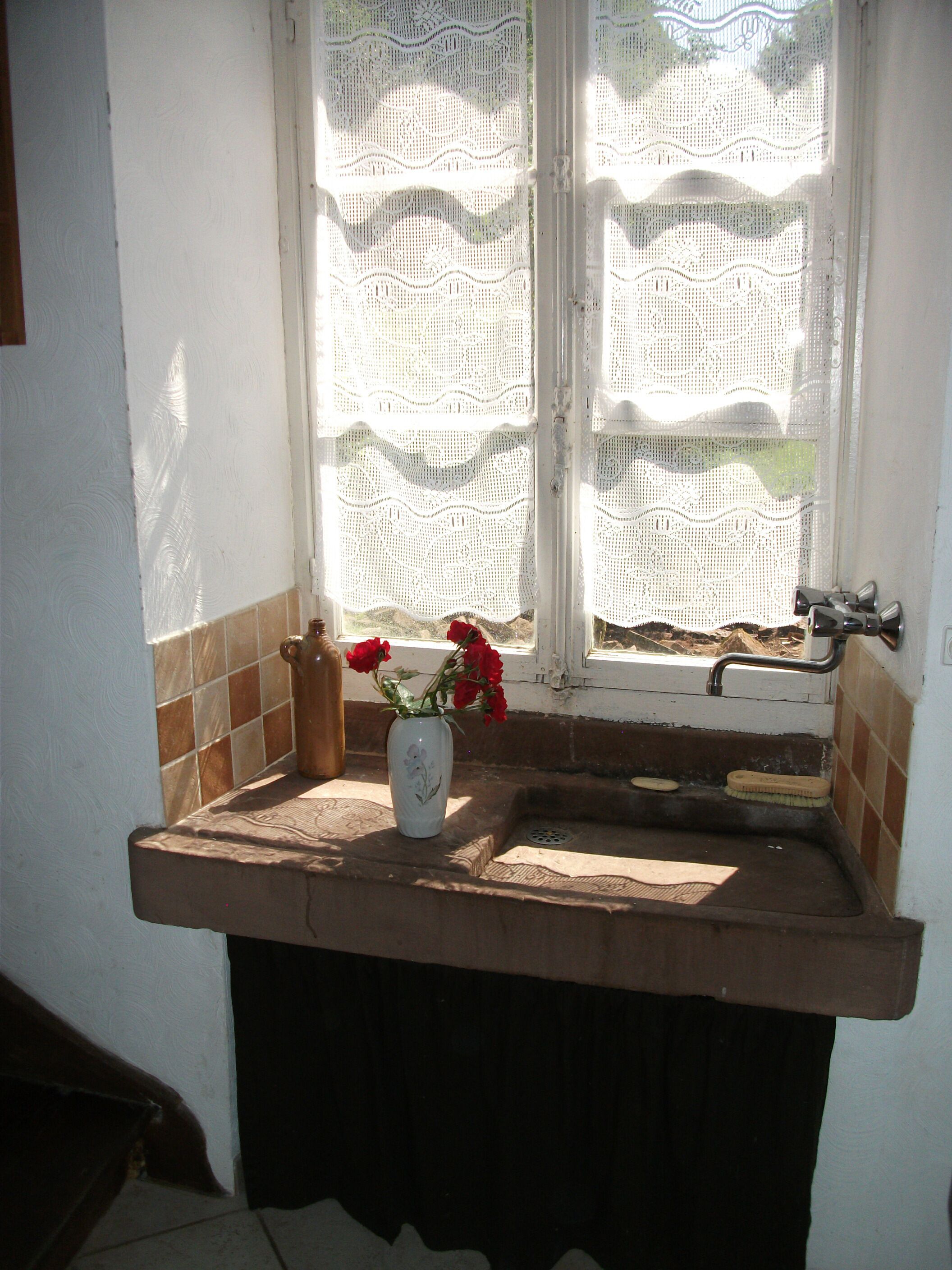 Een badkamer met een wastafel, een raam met kantgordijnen, en een vaas met rode bloemen.