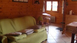 Una habitación acogedora con un sofá verde, una mesita pequeña y una silla. En la pared hay cuadros enmarcados y una ventana con cortinas.