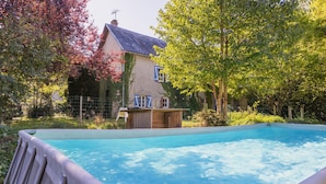 A swimming pool with a wooden deck, a house with blue shutters, and a tree with green leaves.